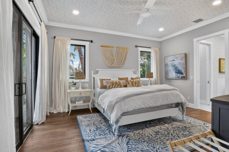 Primary bedroom with white bed, blue patterned rug, macrame wall hanging, heron artwork, black-framed windows, and sliding doors to outdoors.