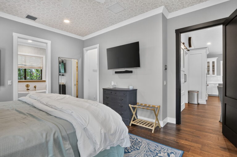 Primary bedroom from foot of bed showing dresser with wall-mounted TV, luggage rack, and open doorways to bathroom and kitchen.