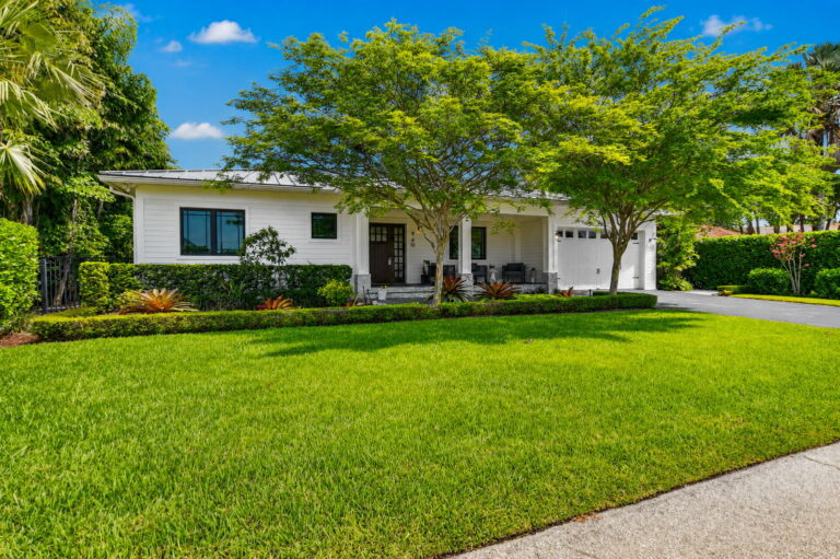 Front exterior with green lawn, landscaped hedge, white home, and shade trees under blue sky.