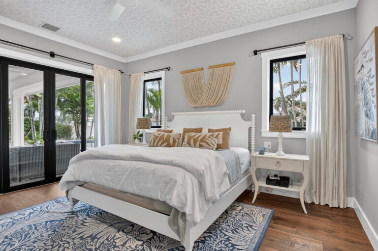 Primary bedroom view toward sliding doors opening to tropical patio, with nightstand, woven lamp, macrame decor, and neutral bedding.