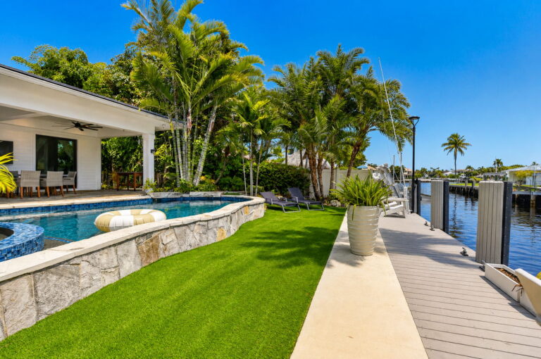 Wide backyard view with pool, stone walkway, covered patio, artificial turf, and long dock along the canal.