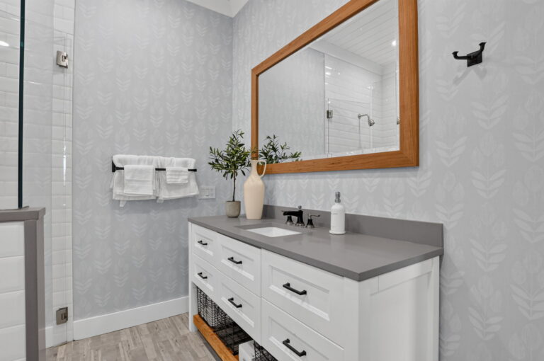 Bathroom vanity with white cabinets, grey countertop, wood-framed mirror, black faucet, towel rack, and matching patterned wallpaper.