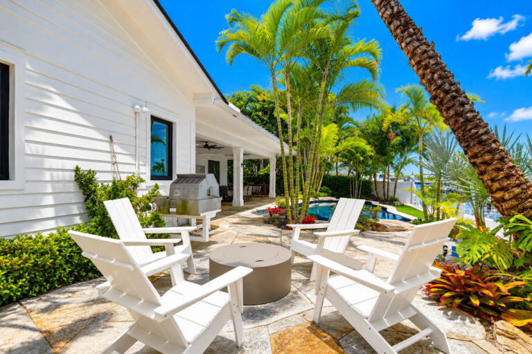 Stone patio with white Adirondack chairs around fire pit table, pool and palms in background, and white home exterior.