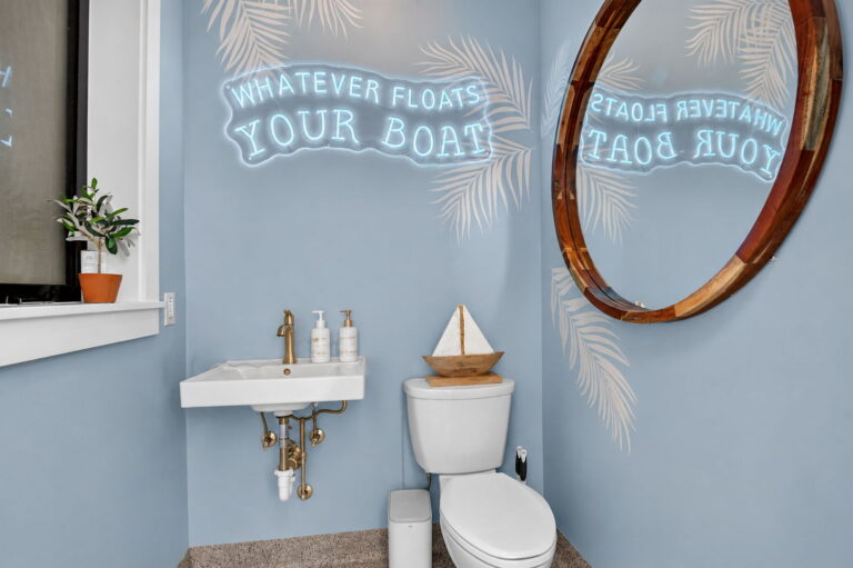 Powder room interior with blue palm-pattern walls, “Whatever Floats Your Boat” neon sign, round wood mirror, wall-hung sink, and sailboat decor.