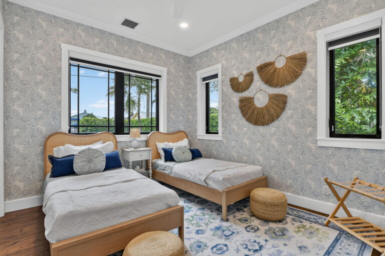 win bedroom with two wood twin beds, tropical wallpaper, woven wall hangings, blue patterned rug, windows to greenery, and woven ottomans.