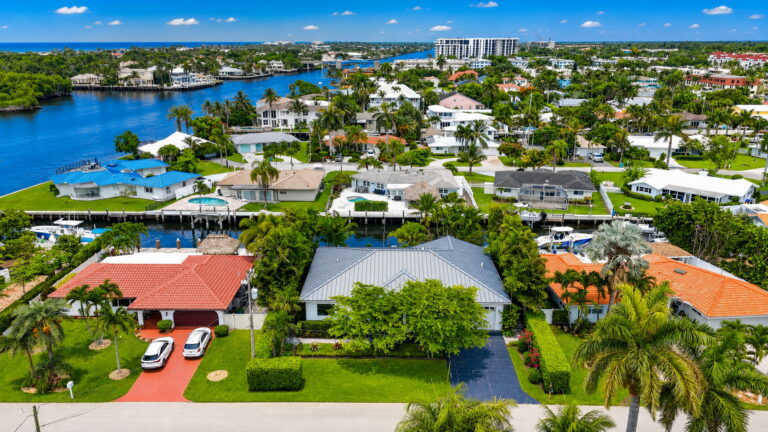 Aerial view of waterfront home with grey roof, landscaped front yard, driveway, canal with docks behind, and neighboring homes.