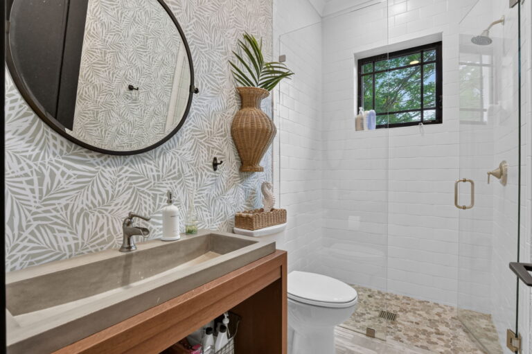Bathroom with wood vanity, grey trough sink, circular mirror, tropical leaf wallpaper, glass walk-in shower, and palm leaf decor.