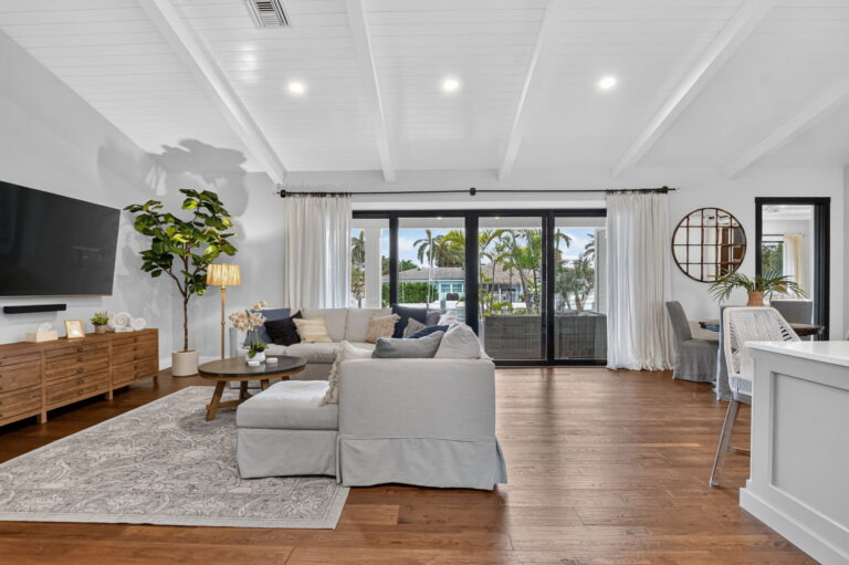 Open living and dining area with TV wall, grey sectional, round dining table with upholstered chairs, circular mirror, and sliding doors to water and palm views.