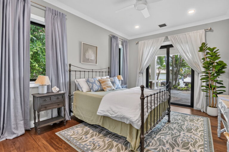 Guest bedroom with metal bed frame, blue floral area rug, nightstand, fiddle-leaf fig, sliding doors to deck, and blue-grey curtains.