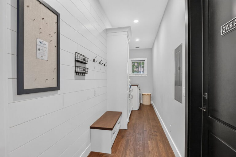 Entry mudroom with shiplap wall, corkboard and hooks, built-in bench with storage, wood floors, garage door on the right, and hallway to laundry room.