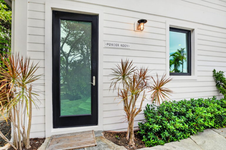 Exterior powder room entrance with black-framed glass door, matching window, outdoor light fixture, and potted tropical plants on white siding.