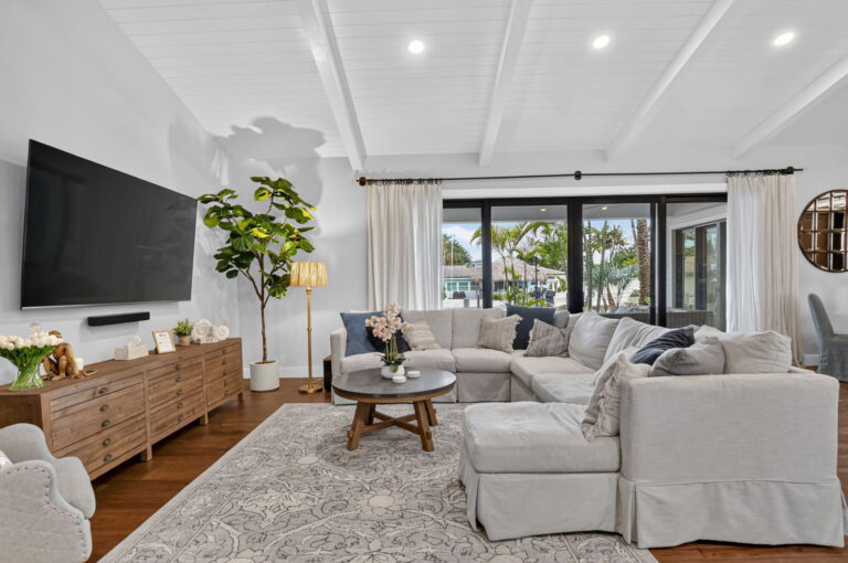 Living room media wall with wall-mounted TV, rustic wood console, fiddle-leaf fig, gold floor lamp, sectional sofa, and sliding doors to tropical outdoor view.