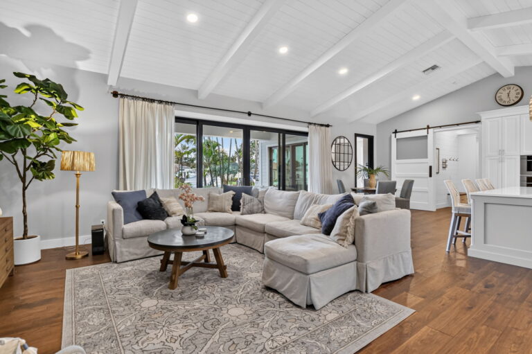 Open living room with vaulted white ceiling, wood beams, grey sectional sofa, round coffee table, patterned rug, and sliding glass doors.