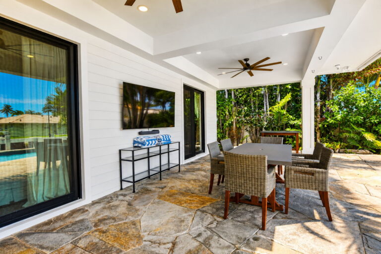 Covered patio dining area with grey table and wicker chairs, wall-mounted TV, shiplap exterior, sliding glass doors, and tropical landscaping.