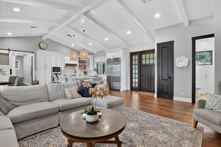 Living room with grey L-shaped sectional, patterned area rug, round wood coffee table, vaulted beam ceiling, and view toward kitchen and black-framed front door.