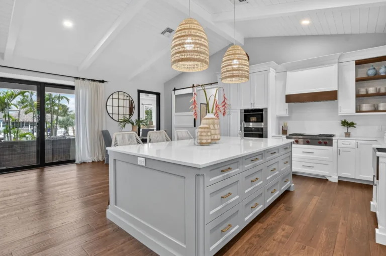 Wide kitchen view with large grey island, white shaker cabinets, woven pendant lights, vaulted beam ceiling, gas range, and open view to patio doors.