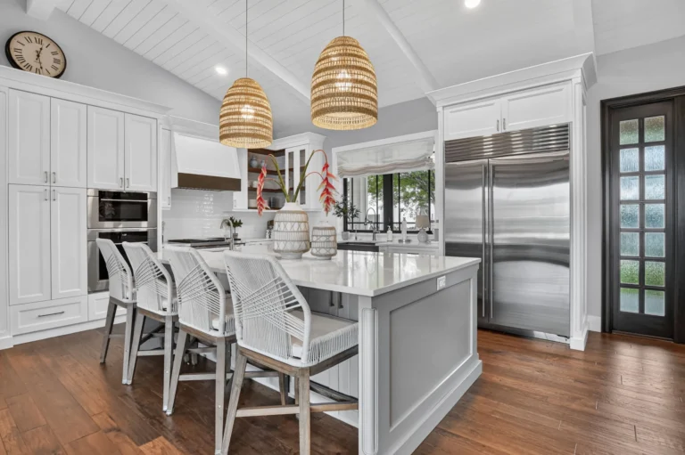 Modern white kitchen with large island, grey woven bar stools, bell-shaped pendant lights, stainless appliances, and wood flooring.