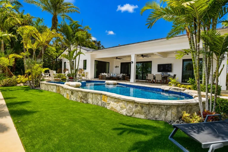 Backyard with curved pool, stone retaining wall, green lawn, outdoor dining and TV, and tropical landscaping.