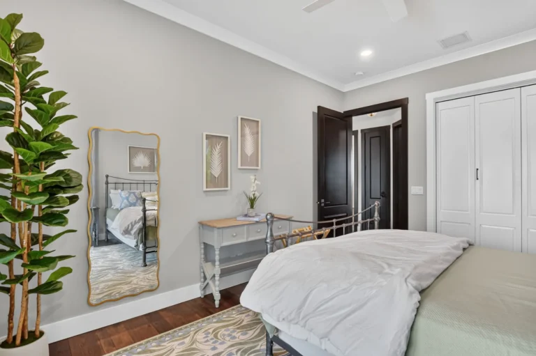 Guest bedroom view with gold arched mirror, grey console table, botanical wall art, open entry door, and white closet doors.