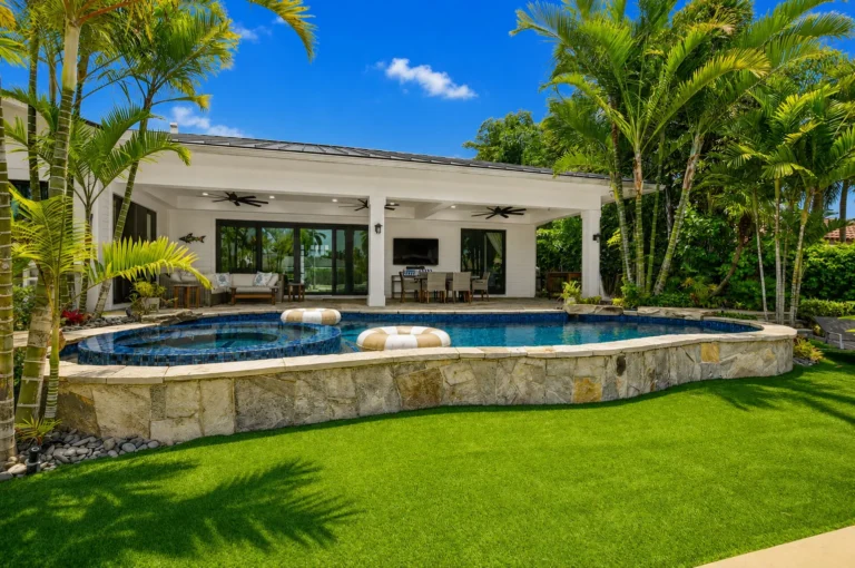 Backyard pool and spa with stone retaining wall, green lawn, covered patio with outdoor seating, and tropical landscaping.