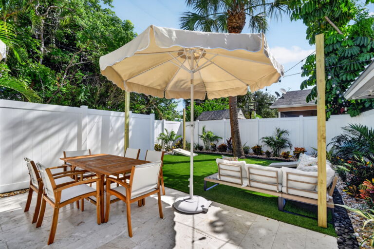Outdoor dining table under a cream umbrella next to an turf lounge area with a built-in sectional and string lights