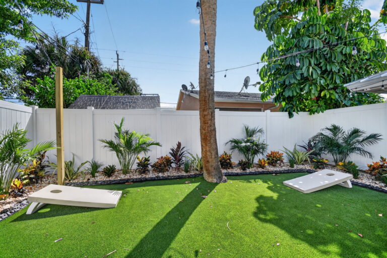 Two cornhole boards on artificial turf lawn, bordered by tropical landscaping, white privacy fence, and string lights