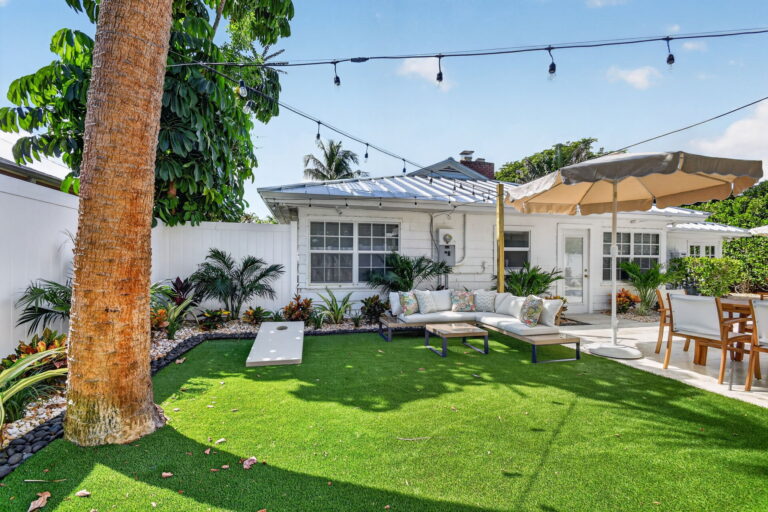 Backyard lawn with cornhole board, white outdoor sectional, dining table, string lights, and the white cottage home in the background