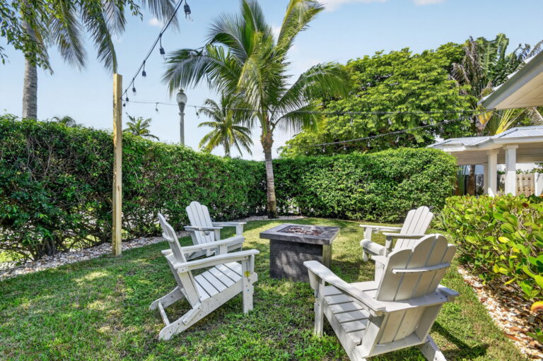 Backyard lawn with square fire pit, white Adirondack chairs, privacy hedge, palm trees, Edison string lights on wooden post, and glimpse of covered patio.
