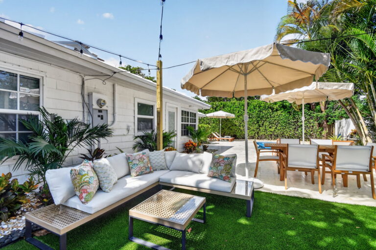 White outdoor sectional sofa with patterned throw pillows on synthetic grass, with dining table, umbrellas, and palm trees in the background