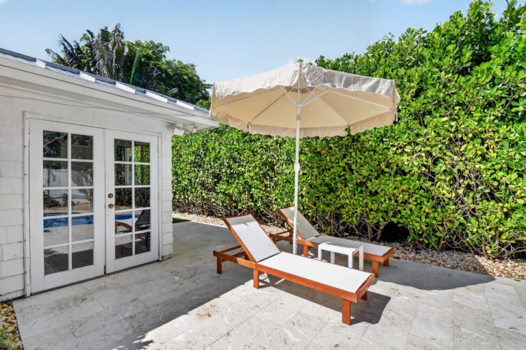 Patio seating area with two wood-and-white lounge chairs, cream umbrella, small side table, and French doors leading into the home
