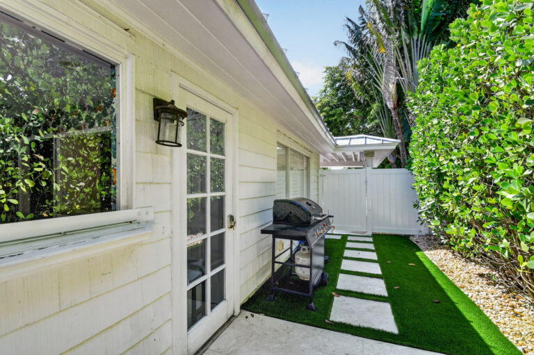Narrow side yard with gas grill, artificial turf pathway with paver stepping stones, white fence gate, and lush greenery