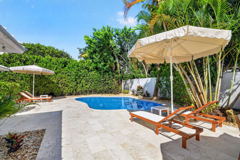Wide view of the backyard pool with multiple lounge chairs, cream umbrellas, potted plants, and travertine pavers