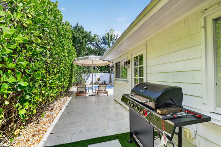 Side patio with black gas grill, tan umbrella and lounge chairs by the pool, tall hedge, and travertine flooring