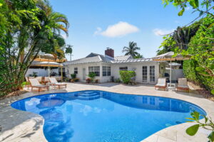 Backyard pool with curved edges, travertine stone deck, wooden lounge chairs, cream umbrellas, and the white cottage-style home framed by palm trees