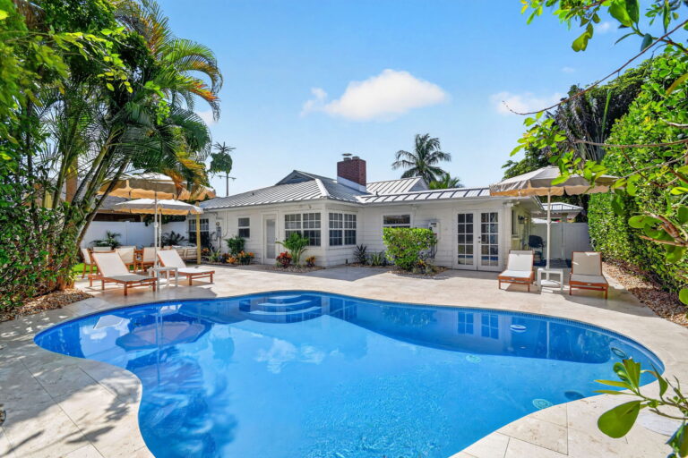 Backyard pool with curved edges, travertine stone deck, wooden lounge chairs, cream umbrellas, and the white cottage-style home framed by palm trees