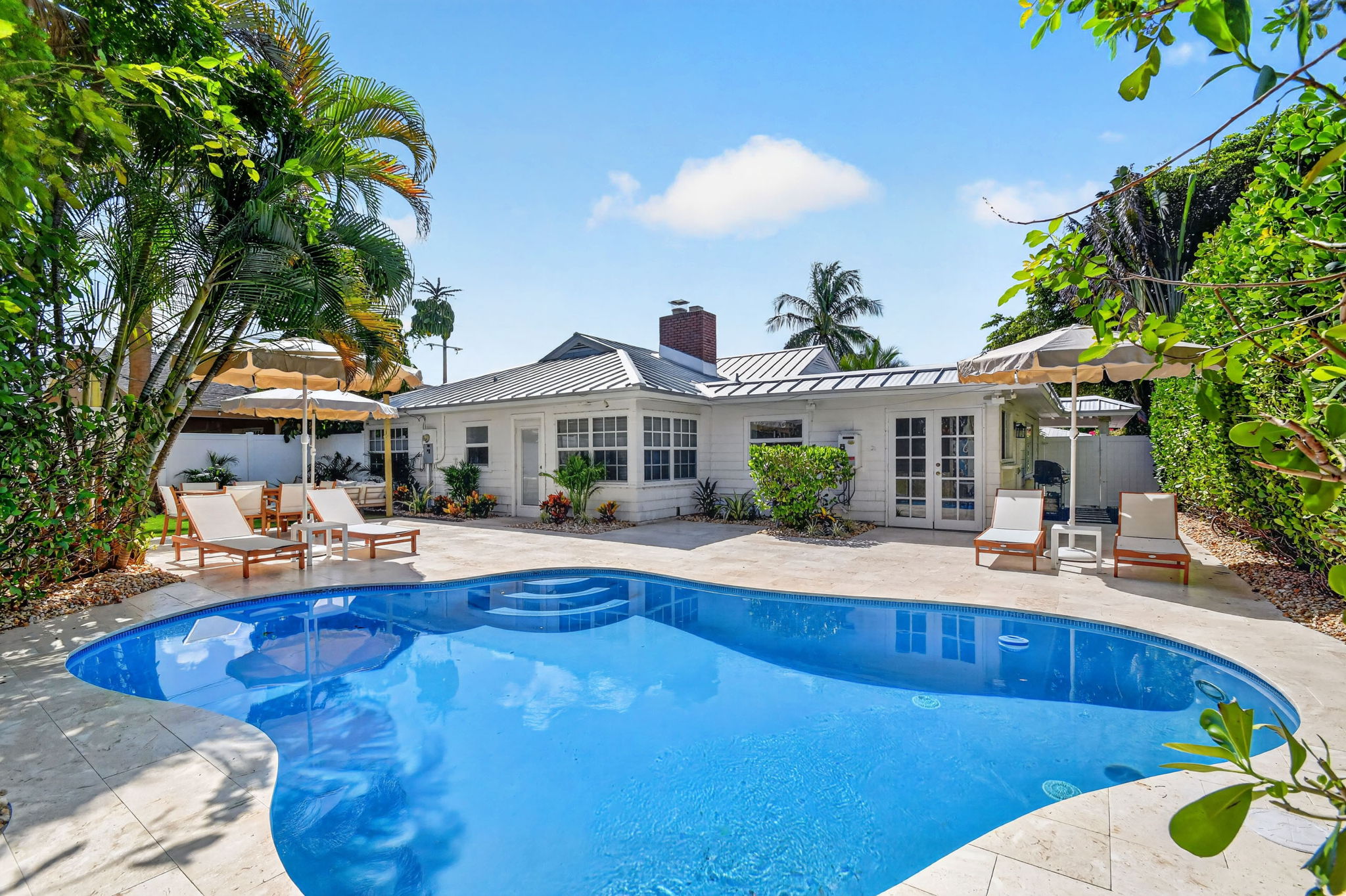 Curved backyard swimming pool with stone steps, lounge chairs, stone patio, and white single-story home under a bright blue sky