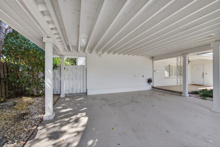 Covered carport with white beam ceiling, support columns, light grey concrete floor, and white picket gate at the rear.